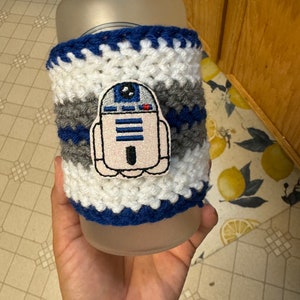 May include: A white, gray, and blue crocheted cup cozy with a white embroidered R2-D2 character.