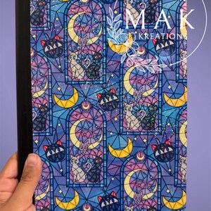 May include: Notebook with a colorful stained-glass design featuring black and white cats, crescent moons, and stars. The cover has a black spine and is held by a hand. The design includes the text "MAK KREATIONS".