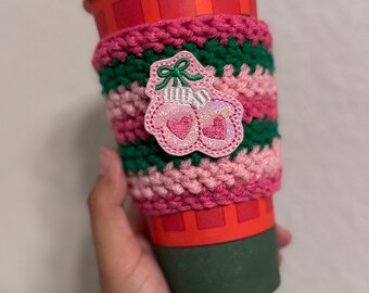 May include: A red paper coffee cup with a white lid, wrapped in a handmade crochet cozy. The cozy features alternating stripes of pink and green yarn, with an embroidered patch of pink ice skates with heart details.