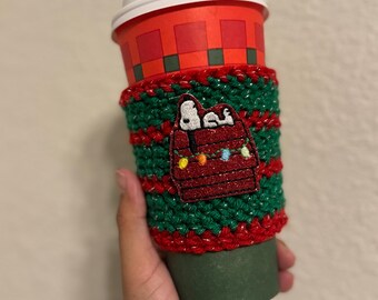 May include: A green and red crocheted coffee cup sleeve with a Snoopy patch. The sleeve is wrapped around a red and white paper coffee cup with a white lid. The Snoopy patch features Snoopy in his doghouse with Christmas lights.