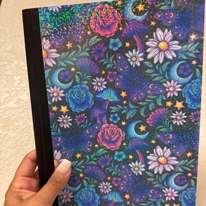 May include: A notebook with a black spine and a colorful cover. The cover features a dark background with a pattern of flowers, moons, mushrooms, and stars in shades of blue, purple, pink, and white. The design has a holographic, iridescent finish.
