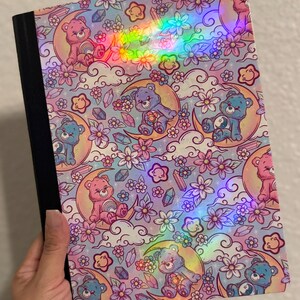 May include: A notebook with a holographic cover featuring a repeating pattern of pink and blue bears sitting on yellow crescent moons, surrounded by white clouds, purple flowers, small stars, and crystals against a light blue background.