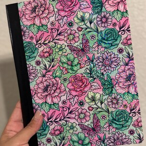 May include: A floral-patterned notebook featuring pink roses, green and teal flowers, and butterflies on a pink and light green background. The notebook has a black spine and is held by a hand.