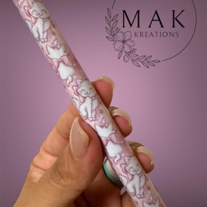 May include: A pink and white pen featuring a repeating pattern of white cartoon cats with pink bows. The pen has a pink tip and a silver metal end. The pen is held against a soft purple background.