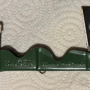 May include: A vintage Singer Sewing Machines attachment tool featuring a dark green metal body with a scalloped top edge. The device includes a silver-toned metal component on the left and a black and silver-toned metal blade on the right side.