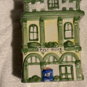 May include: A ceramic building figurine painted in shades of green, yellow, and white. The structure features a gabled roof, arched windows, and a sign that reads POST OFFICE. A small blue mailbox with the word MAIL is positioned at the base.