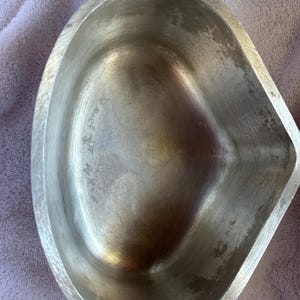 May include: A vintage, oval-shaped metal serving dish with a handle. The dish has a textured handle and a smooth, slightly worn interior. The metal has a silver color with some signs of age and use, adding to its antique charm.