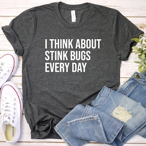 May include: A dark gray t-shirt with white text that reads "I think about stink bugs every day". The shirt is laid flat on a white wooden surface with a pair of white sneakers, blue jeans, and a potted plant.