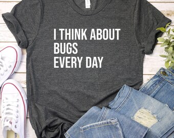 May include: A dark grey t-shirt with white text that reads "I think about bugs every day". The shirt is laid flat on a white wooden surface with a pair of white sneakers and a pair of blue jeans.