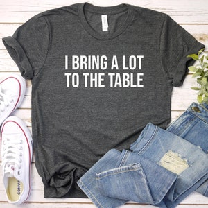 May include: A dark gray t-shirt with the text "I bring a lot to the table" printed in white on the front. The shirt is laid flat on a white wooden surface with a pair of white sneakers and blue jeans.