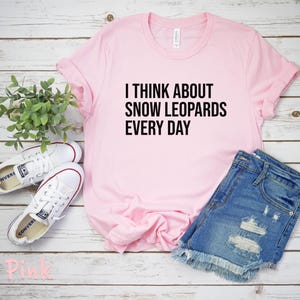 May include: A light pink t-shirt with the text "I THINK ABOUT SNOW LEOPARDS EVERY DAY" in black. The shirt is paired with denim shorts and white sneakers. The items are arranged on a light wood-grain surface.