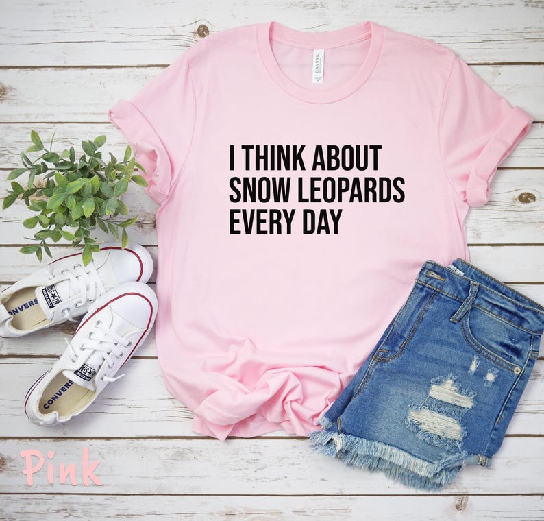 May include: A light pink t-shirt with the text "I THINK ABOUT SNOW LEOPARDS EVERY DAY" in black. The shirt is paired with denim shorts and white sneakers. The items are arranged on a light wood-grain surface.
