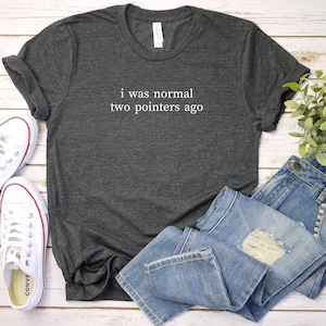 May include: Dark gray t-shirt with the text "i was normal two pointers ago" in white. The shirt is paired with light blue denim jeans and white sneakers with red accents. A sprig of greenery is also present.