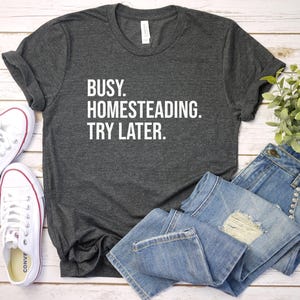 May include: Dark gray t-shirt with the text "BUSY. HOMESTEADING. TRY LATER." White canvas sneakers and blue jeans are arranged on a light wood surface. A small green plant is also visible.
