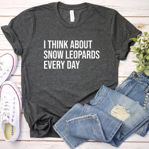 May include: Dark gray t-shirt with the text "I THINK ABOUT SNOW LEOPARDS EVERY DAY" in white. The shirt is paired with blue jeans and white sneakers with red accents. A small green plant is also in the image.