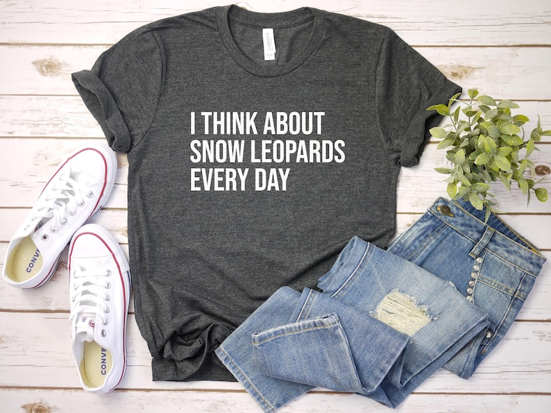 May include: Dark gray t-shirt with the text "I THINK ABOUT SNOW LEOPARDS EVERY DAY" in white. The shirt is paired with blue jeans and white sneakers with red accents. A small green plant is also in the image.
