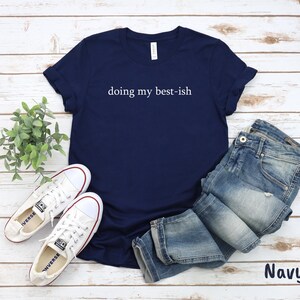 May include: Navy blue t-shirt with the text "doing my best-ish" in white. The shirt is paired with blue jeans and white canvas sneakers. The word "Navy" is displayed in the bottom right corner.