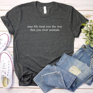 May include: Charcoal grey t-shirt with the white text "may life treat you the way that you treat animals". The image includes blue denim jeans, white trainers, and a small plant, all on a light wood surface.