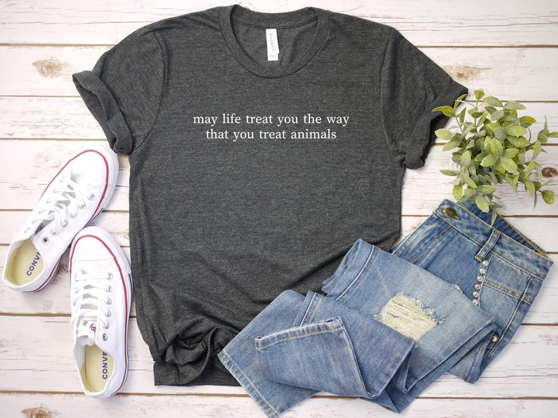 May include: Charcoal grey t-shirt with the white text "may life treat you the way that you treat animals". The image includes blue denim jeans, white trainers, and a small plant, all on a light wood surface.