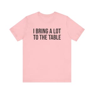 Może przedstawiać: R&oacute;żowy t-shirt z nadrukiem "I bring a lot to the table" w kolorze czarnym.