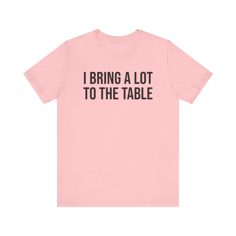 Może przedstawiać: R&oacute;żowy t-shirt z nadrukiem "I bring a lot to the table" w kolorze czarnym.