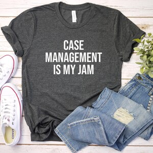 May include: Dark gray t-shirt with the text "CASE MANAGEMENT IS MY JAM" in white. The shirt is paired with light blue denim jeans and white canvas sneakers. A small green plant is also in the image.