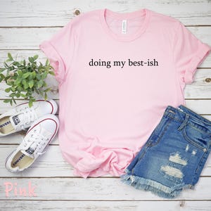 May include: A light pink t-shirt with the phrase "doing my best-ish" in black lettering. The shirt is paired with denim shorts and white sneakers. The items are arranged on a light wood-grain surface.