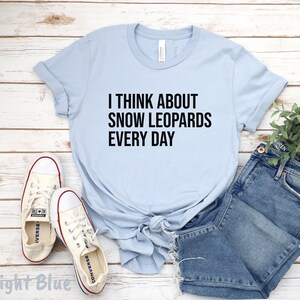 May include: Light blue t-shirt with the text "I THINK ABOUT SNOW LEOPARDS EVERY DAY" in black. The shirt is paired with blue denim shorts and white sneakers with red accents. The shirt is knotted at the waist.