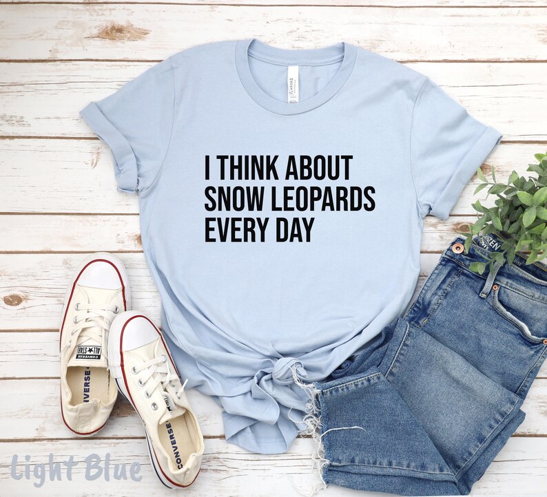 May include: Light blue t-shirt with the text "I THINK ABOUT SNOW LEOPARDS EVERY DAY" in black. The shirt is paired with blue denim shorts and white sneakers with red accents. The shirt is knotted at the waist.