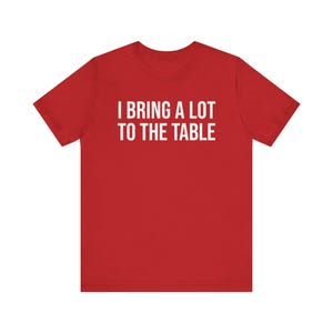 Może przedstawiać: Czerwony T-shirt z kr&oacute;tkim rękawem z białym napisem "Wnoszę wiele do stołu."