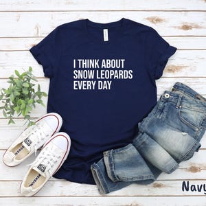 May include: Navy blue t-shirt with the text "I THINK ABOUT SNOW LEOPARDS EVERY DAY" in white. The shirt is paired with blue jeans and white sneakers. The word "Navy" is printed in the bottom right corner.