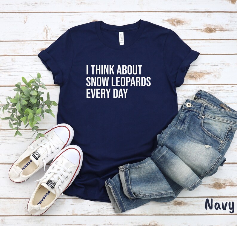 May include: Navy blue t-shirt with the text "I THINK ABOUT SNOW LEOPARDS EVERY DAY" in white. The shirt is paired with blue jeans and white sneakers. The word "Navy" is printed in the bottom right corner.
