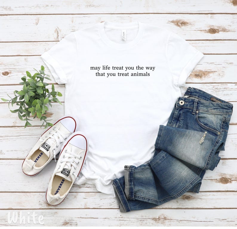 May include: A white t-shirt with the text "may life treat you the way that you treat animals." The image also shows blue denim jeans and white canvas trainers with red detailing. A small green plant is also in the image.