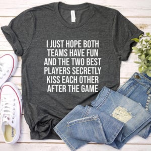 May include: Dark gray t-shirt with the text "I JUST HOPE BOTH TEAMS HAVE FUN AND THE TWO BEST PLAYERS SECRETLY KISS EACH OTHER AFTER THE GAME." White sneakers and blue jeans are also in the image.