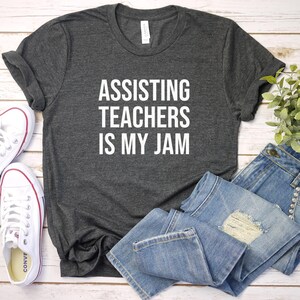 May include: Charcoal grey t-shirt with the slogan "ASSISTING TEACHERS IS MY JAM" in white. The image includes light blue denim jeans and white trainers. A small green plant is also visible.