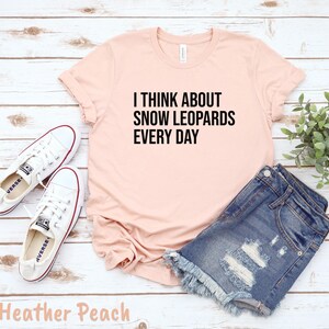 May include: A heather peach t-shirt with the text "I THINK ABOUT SNOW LEOPARDS EVERY DAY" in black. The shirt is paired with denim shorts and white sneakers. The outfit is laid out on a light wood surface.