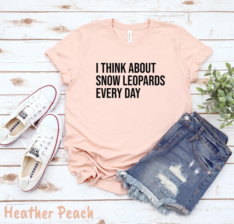 May include: A heather peach t-shirt with the text "I THINK ABOUT SNOW LEOPARDS EVERY DAY" in black. The shirt is paired with denim shorts and white sneakers. The outfit is laid out on a light wood surface.