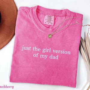 Może przedstawiać: Jaskraworóżowy t-shirt z krótkim rękawem, okrągłym dekoltem i białym napisem "just the girl version of my dad" z przodu. Koszulka posiada metkę marki Comfort Colors po wewnętrznej stronie kołnierza, wskazującą na rozmiar S.