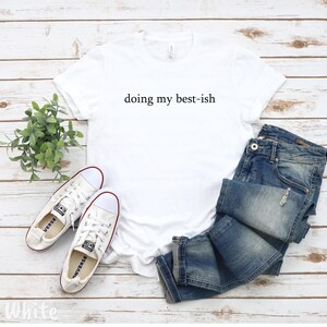 May include: A white t-shirt with the phrase "doing my best-ish" printed in black, paired with blue jeans and white sneakers with red accents. The casual outfit is arranged on a light wood-grain surface.