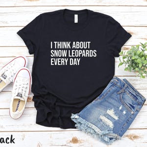 May include: Black t-shirt with the text "I THINK ABOUT SNOW LEOPARDS EVERY DAY" in white. The shirt is paired with denim shorts and white sneakers. A small green plant is also in the image.