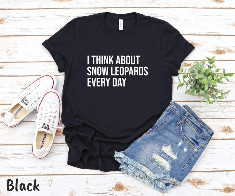 May include: Black t-shirt with the text "I THINK ABOUT SNOW LEOPARDS EVERY DAY" in white. The shirt is paired with denim shorts and white sneakers. A small green plant is also in the image.