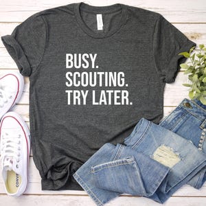 May include: Dark gray t-shirt with the text "BUSY. SCOUTING. TRY LATER." White canvas sneakers and blue jeans are also in the image. The shirt is folded and placed on a light wood surface.