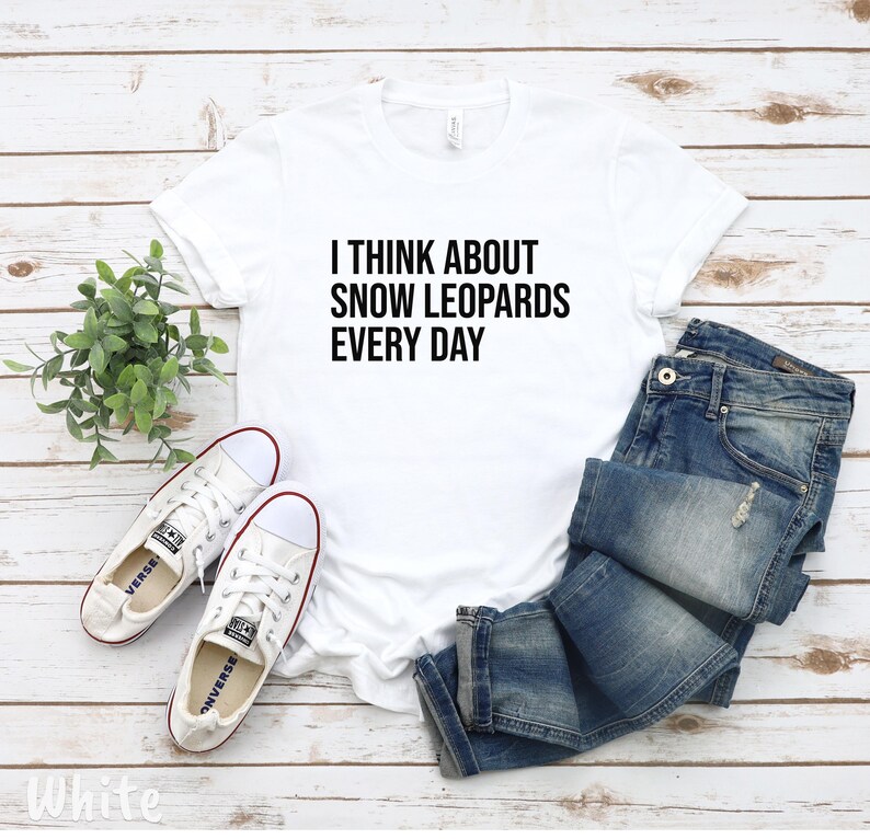 May include: White t-shirt with the text "I THINK ABOUT SNOW LEOPARDS EVERY DAY" in black. The shirt is paired with blue jeans and white sneakers with red accents. A small green plant is also in the image.