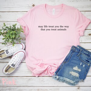 May include: A pale pink t-shirt with the message "may life treat you the way that you treat animals." The image also shows denim shorts and white trainers. The shirt is laid on a white wooden surface.