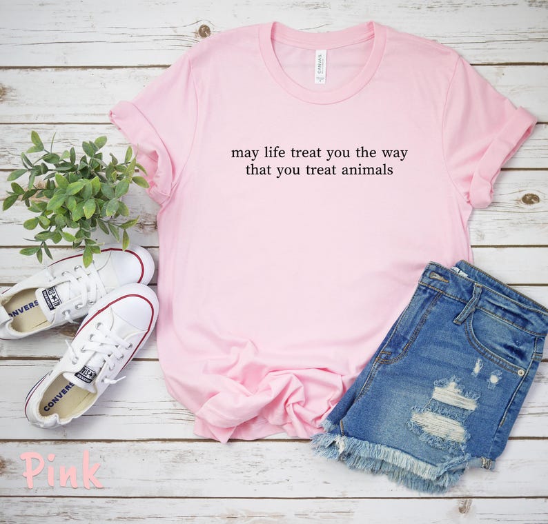 May include: A pale pink t-shirt with the message "may life treat you the way that you treat animals." The image also shows denim shorts and white trainers. The shirt is laid on a white wooden surface.