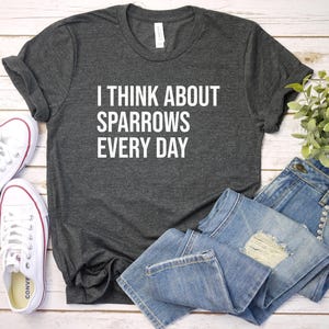 May include: A charcoal grey t-shirt with the white text "I THINK ABOUT SPARROWS EVERY DAY". The image also shows blue denim jeans and white trainers with red detailing, set on a light wooden surface.