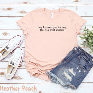 May include: A peach-coloured t-shirt with the text "may life treat you the way that you treat animals" in black. The shirt is paired with denim shorts and white trainers. The words "Heather Peach" are at the bottom.