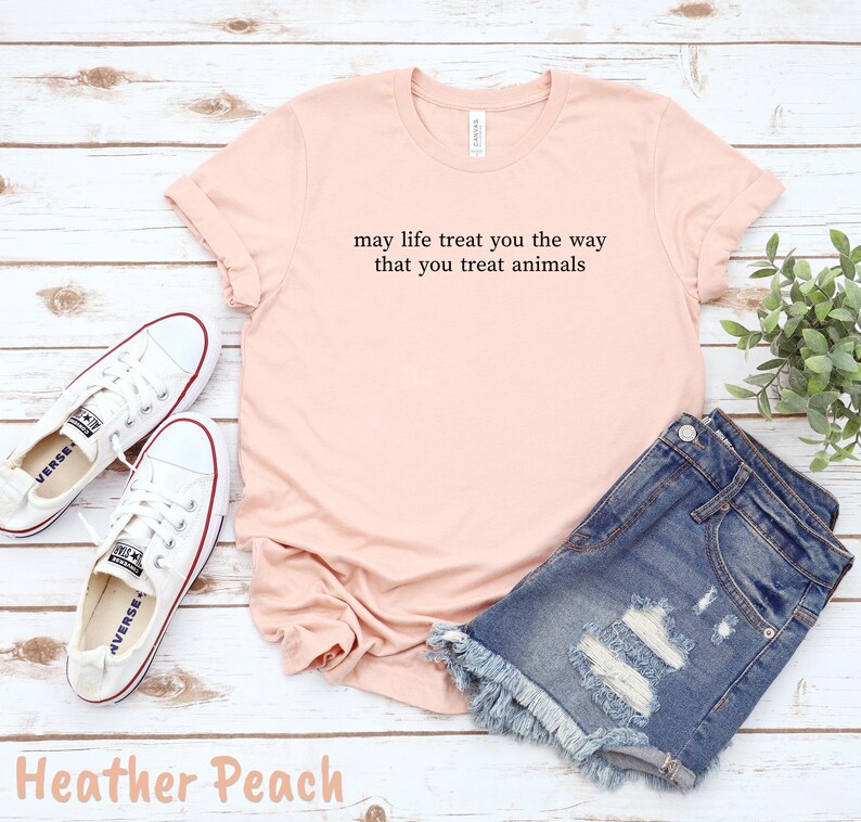 May include: A peach-coloured t-shirt with the text "may life treat you the way that you treat animals" in black. The shirt is paired with denim shorts and white trainers. The words "Heather Peach" are at the bottom.