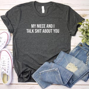 Może przedstawiać: Ciemnoszary melanżowy t-shirt z krótkim rękawem z białym napisem "MY NIECE AND I TALK SHIT ABOUT YOU" z przodu. Koszulka jest pokazana obok pary białych tenisówek z płótna oraz niebieskich dżinsów z przetarciami.