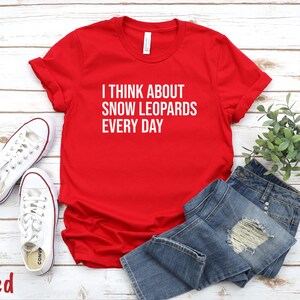 May include: A red t-shirt with the text "I THINK ABOUT SNOW LEOPARDS EVERY DAY" in white. The shirt is paired with blue jeans and white sneakers. The word "Red" is at the bottom left.
